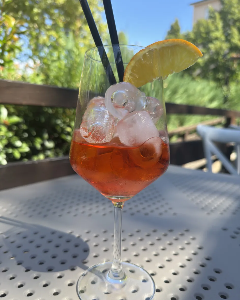 Uno spritz con ghiaccio e fetta d'arancia sul tavolo all'aperto de La Vecchia Pesa, con il verde del parco di Cassino dietro