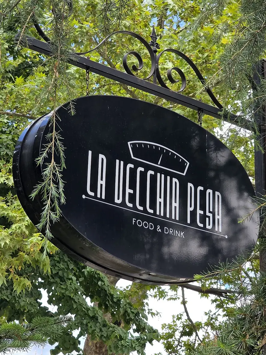 L'insegna ovale «La Vecchia Pesa — Food & Drink» appesa tra gli alberi del parco