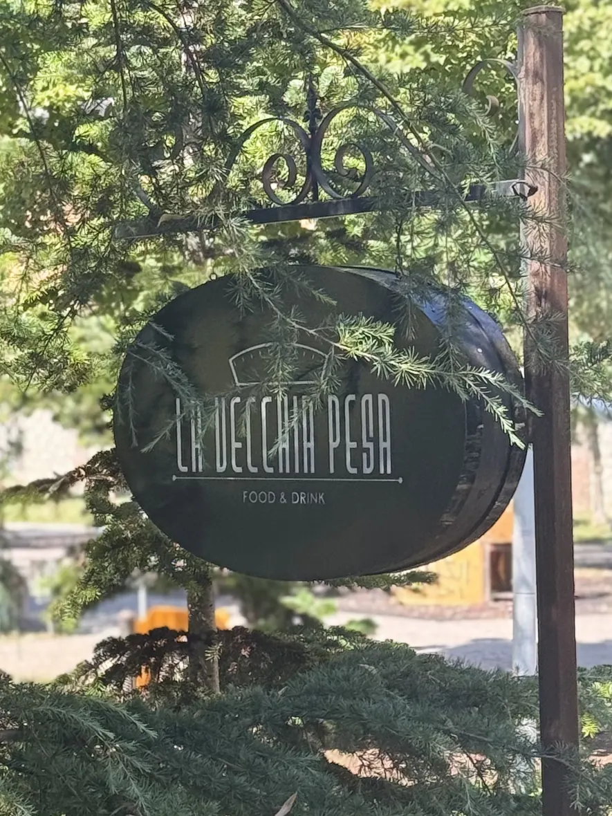 L'insegna ovale «La Vecchia Pesa — Food & Drink» appesa al palo in ferro battuto, fra i rami dei cedri del parco