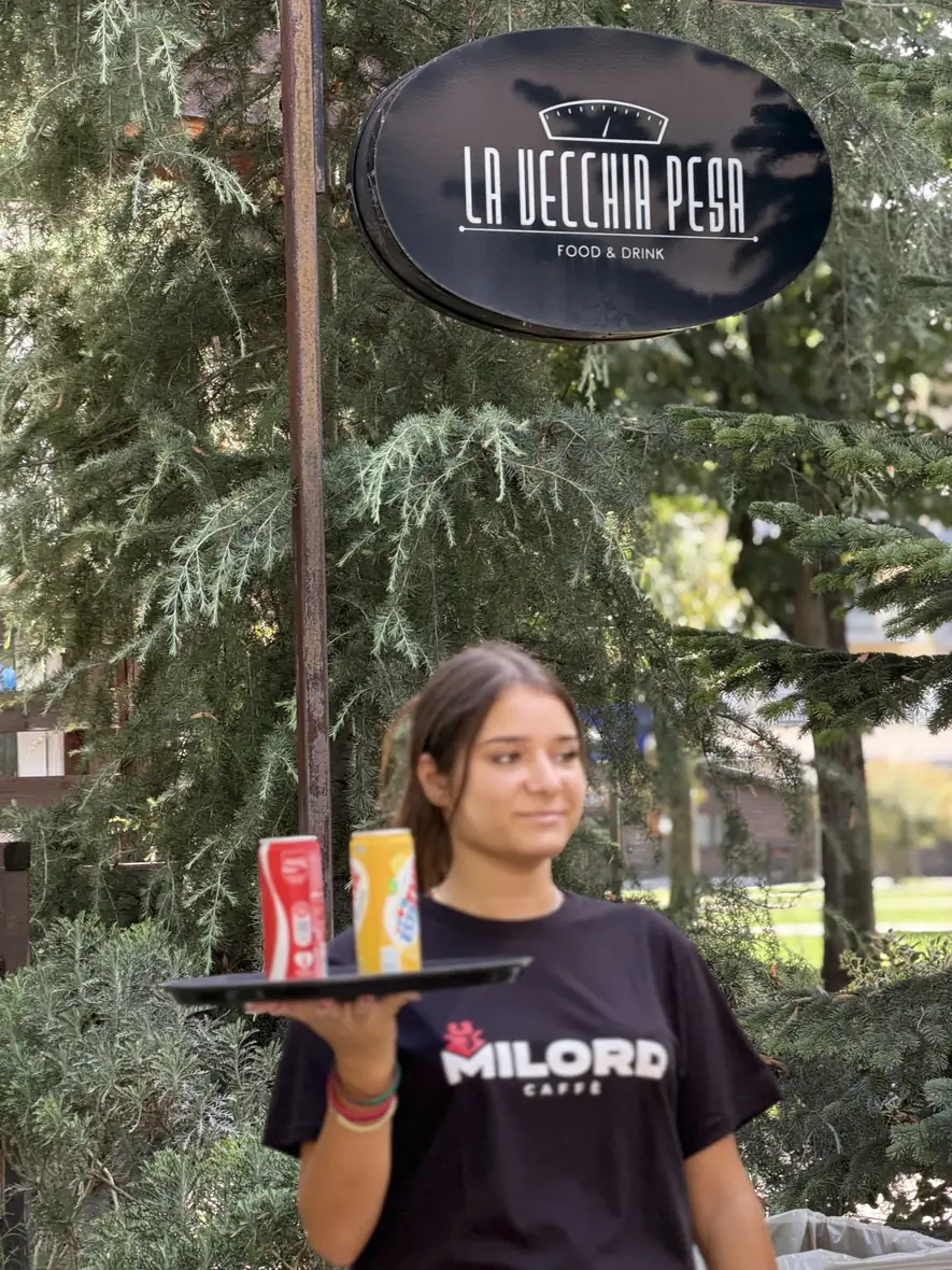 Una cameriera de La Vecchia Pesa con il vassoio, sotto l'insegna ovale appesa fra gli alberi