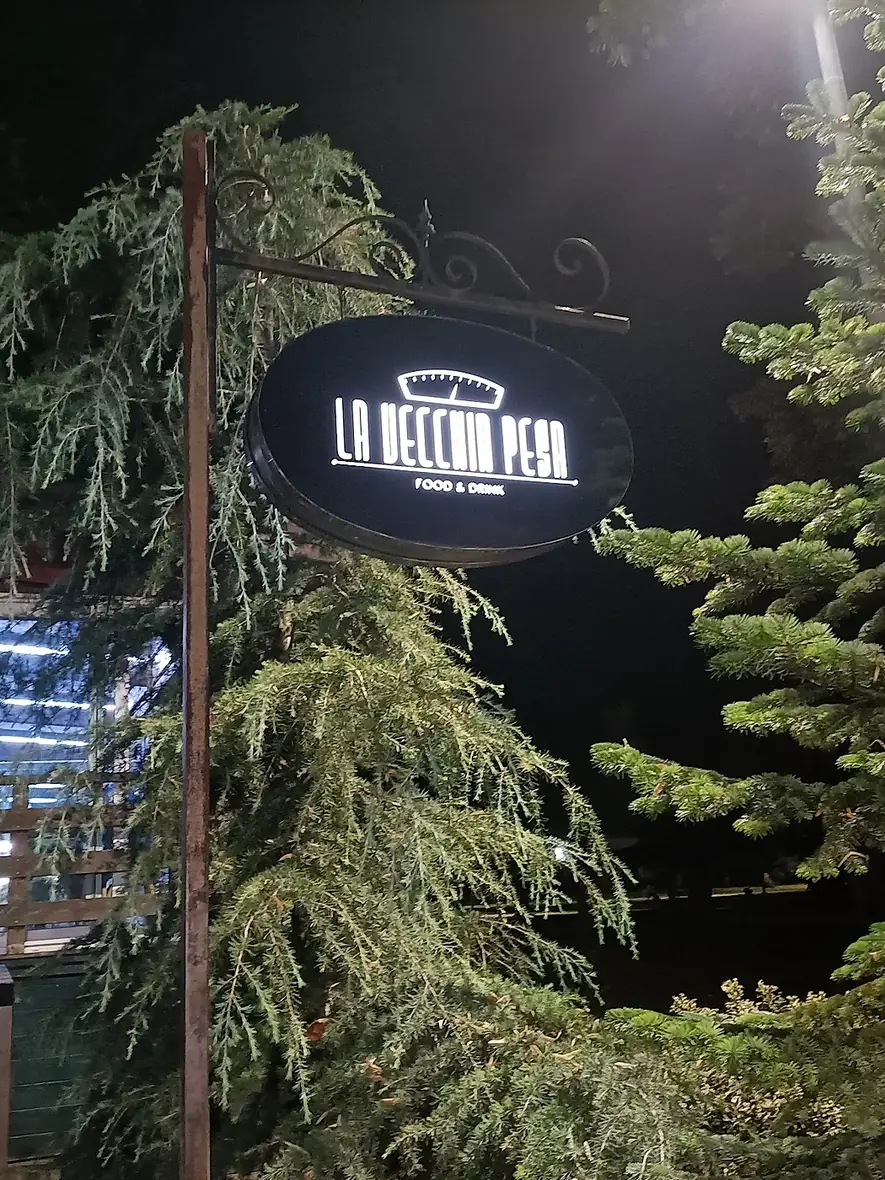 L'insegna ovale de La Vecchia Pesa accesa di sera fra i rami dei cedri, con le luci della veranda dietro gli alberi del parco